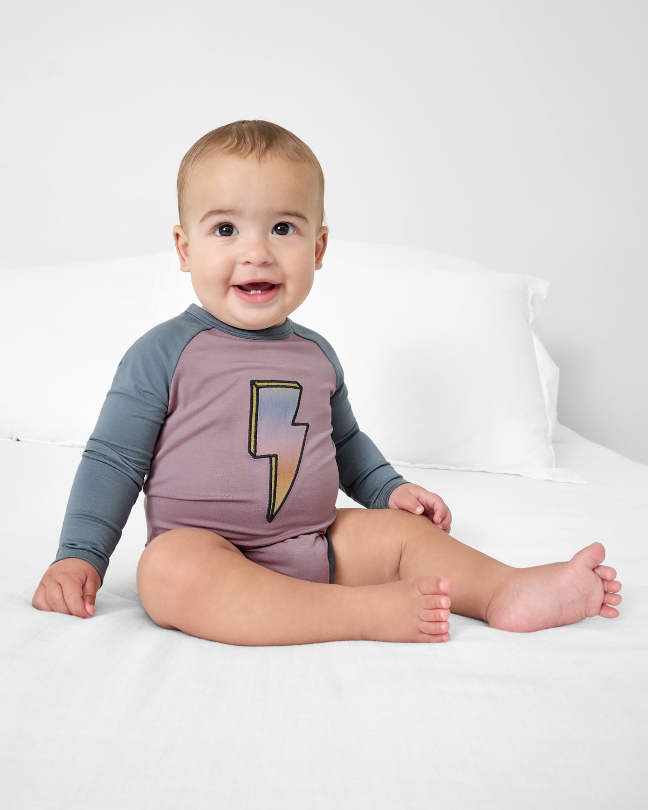 Baby sitting on a white bed in the Bolt Raglan Bodysuit