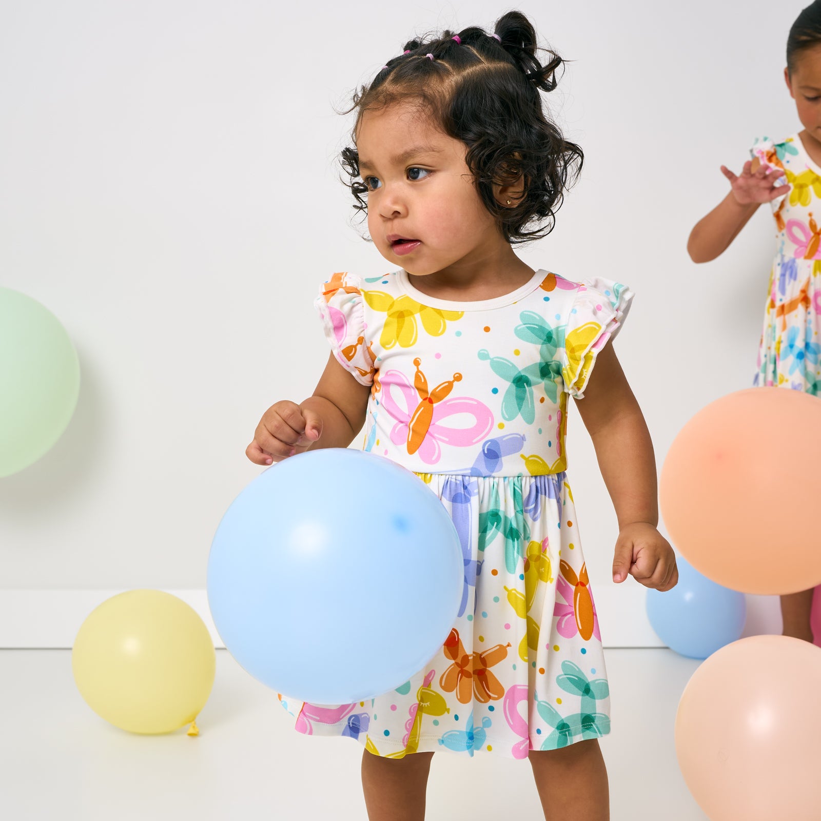 Standing child wearing the Balloon Bash Flutter Twirl Dress with Bodysuit with balloons on the floor