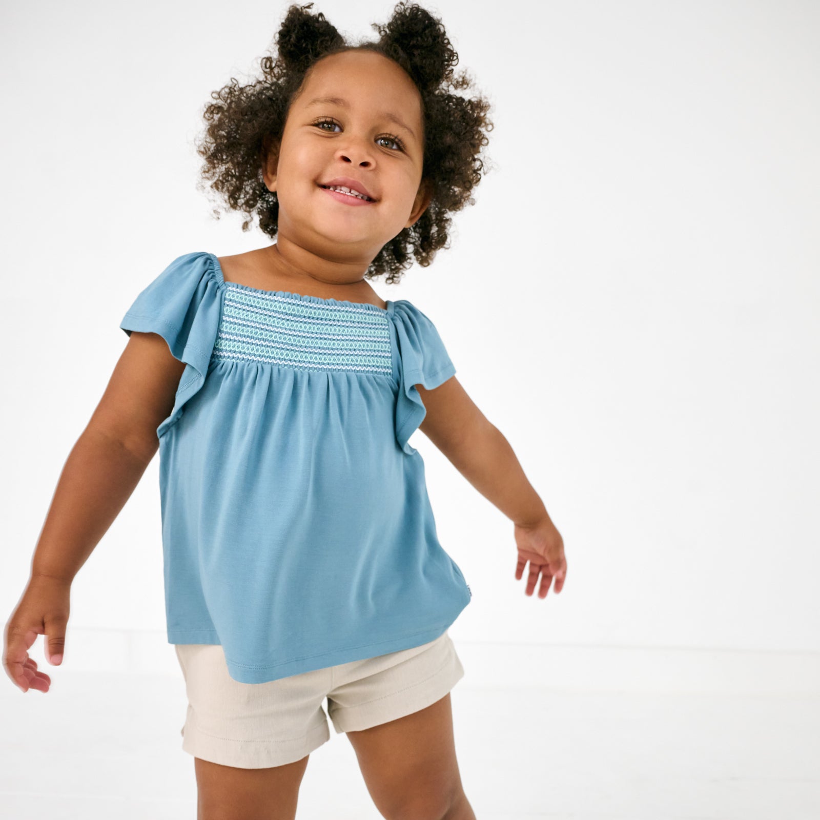 Standing child wearing the Dusk Blue Smocked Flutter Top with denim shorts