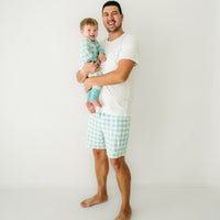 Man holding his child. Dad is wearing Aqua Gingham men's pajama shorts paired with a men's Bright White short sleeve pajama top. His child is wearing a matching Aqua Gingham zippy