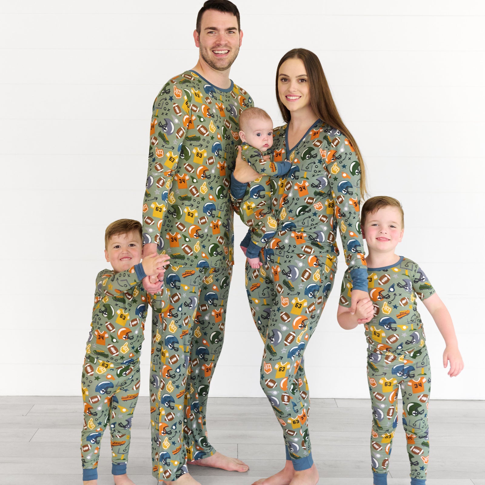 Family in the Touchdown Time collection. Boy on the left is in Touchdown Time Two-piece Pajama Set and holding his father's hand, who is in the touchdown-time-mens-pajama-top & pants. Baby in the middle is in the Touchdown Time Zippy, being held by mother who is in the touchdown-time-womens-pajama-top & pants. Boy on the right is in the touchdown-time-two-piece-short-sleeve-pajama-set