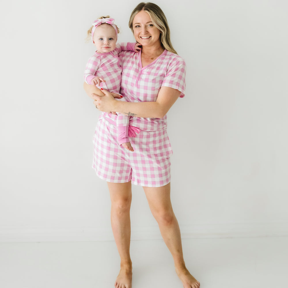 Woman holding her child wearing matching Pink Gingham pajamas. Mom is wearing women's Pink Gingham pajama shorts and matching women's short sleeve pajama top. Child is wearing a matching Pink Gingham zippy paired with a matching luxe bow headband 
