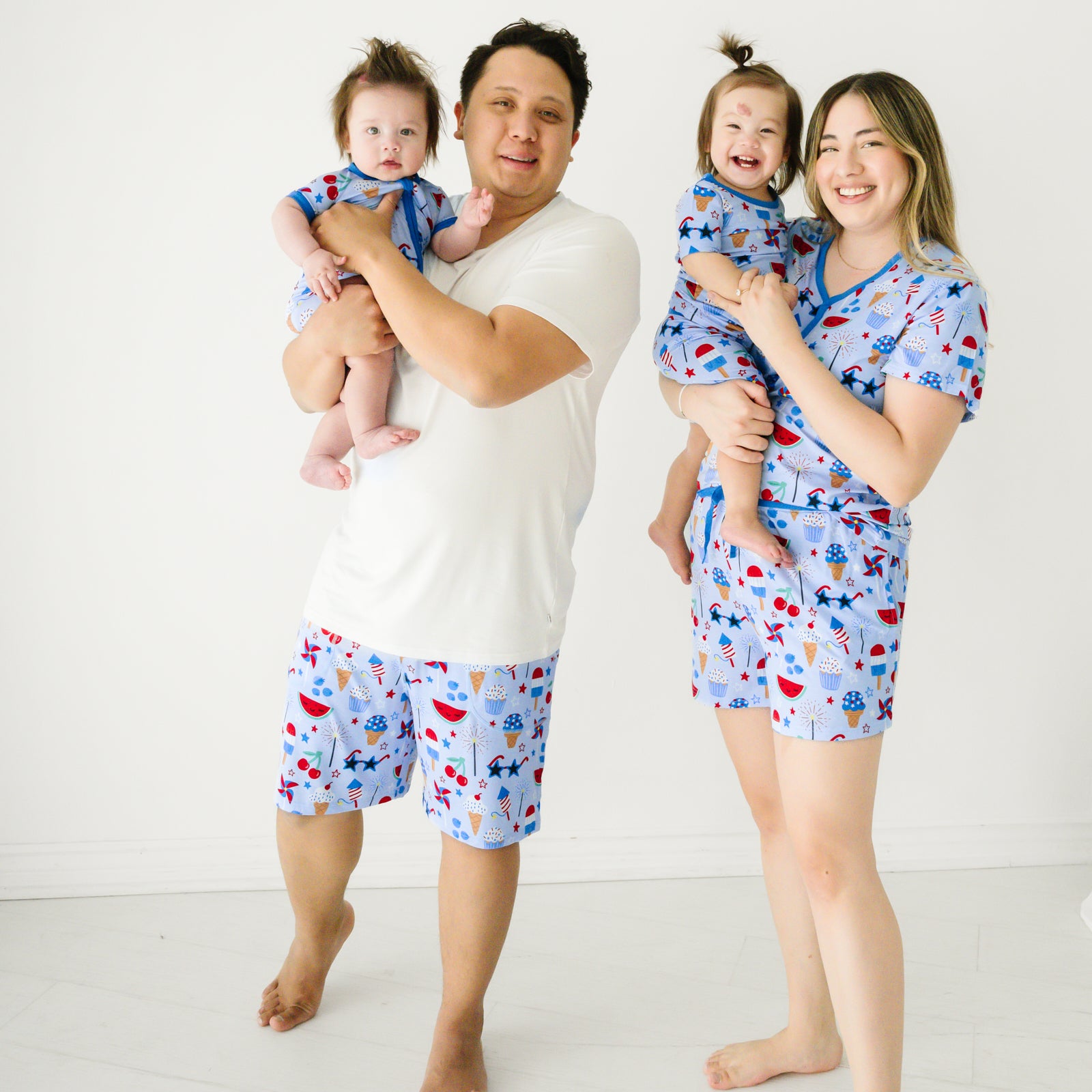 Family of four wearing matching Stars, Stripes, and Sweets pajamas. Dad is wearing a bright white men's pj top paired with men's Stars, Stripes, and Sweets men's pj shorts. Mom is wearing a Stars and Stripes and Sweets women's pj top and matching women's pj shorts. Their kids are wearing Stars, Stripes, and Sweets in zippy and two piece styles