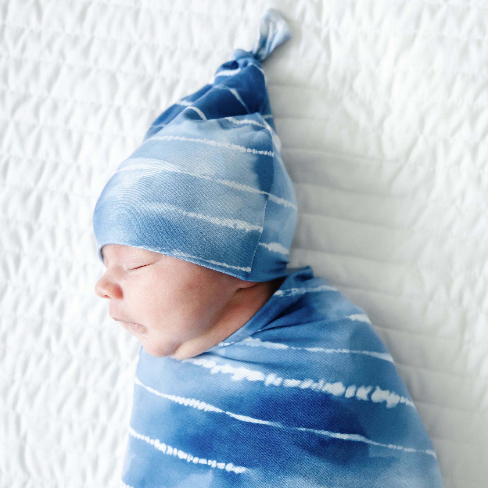 Close up image of an infant swaddled in a Blue Tie Dye Dreams swaddle & hat set