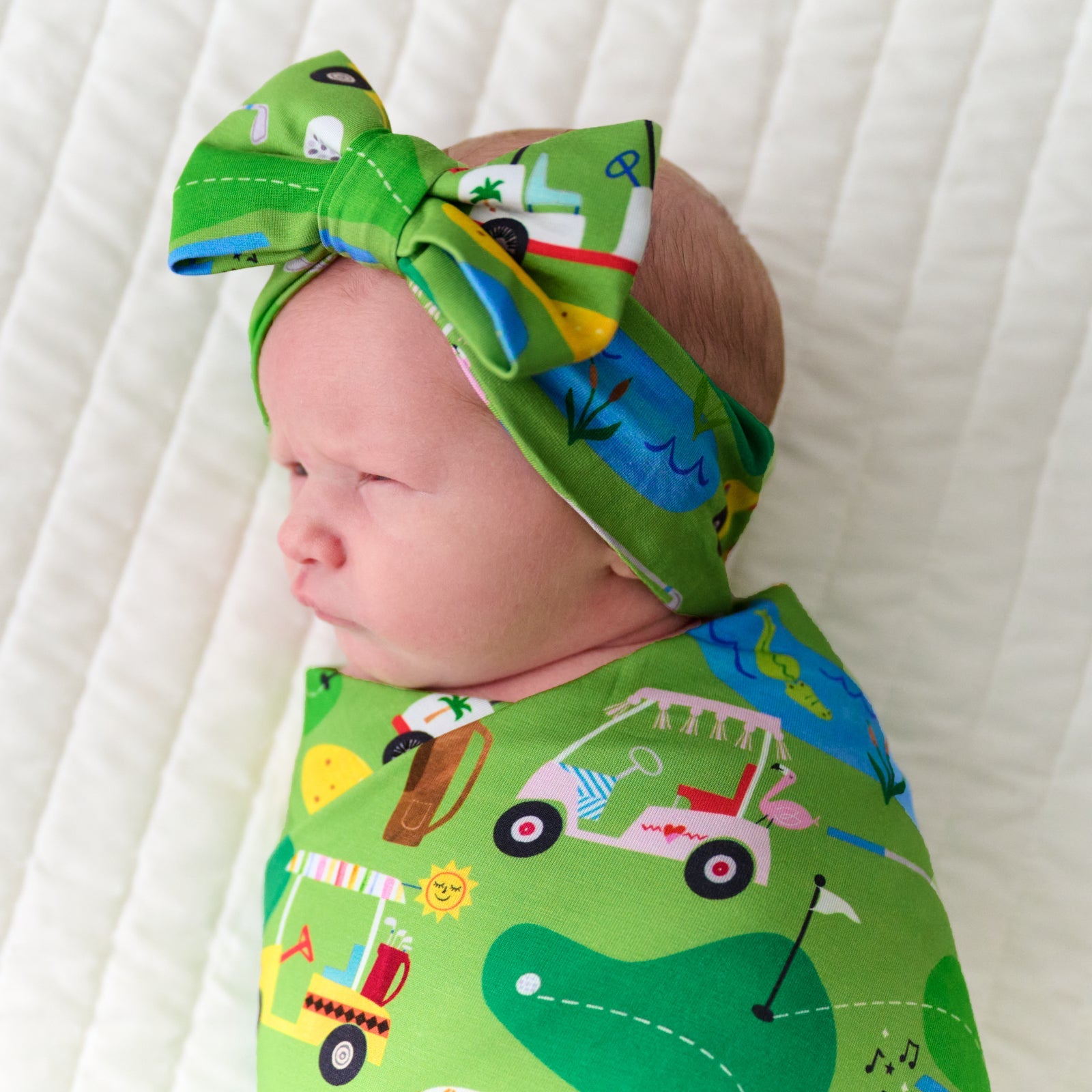 Close up image of an infant swaddled in a Fairway Fun swaddle and luxe bow headband set