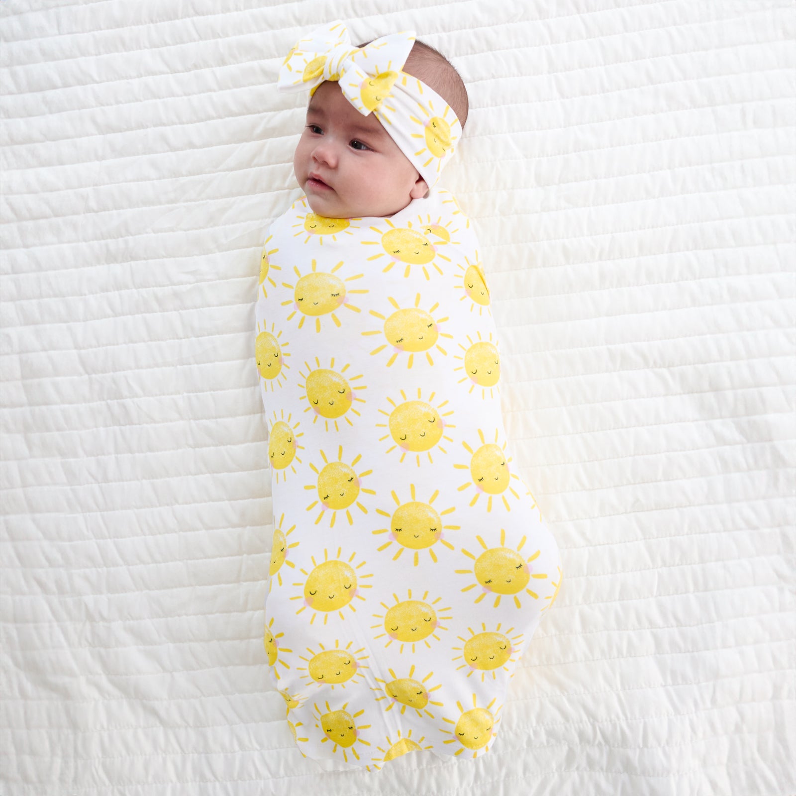 Child laying on a bed swaddled in a Sunshine swaddle and luxe bow headband set