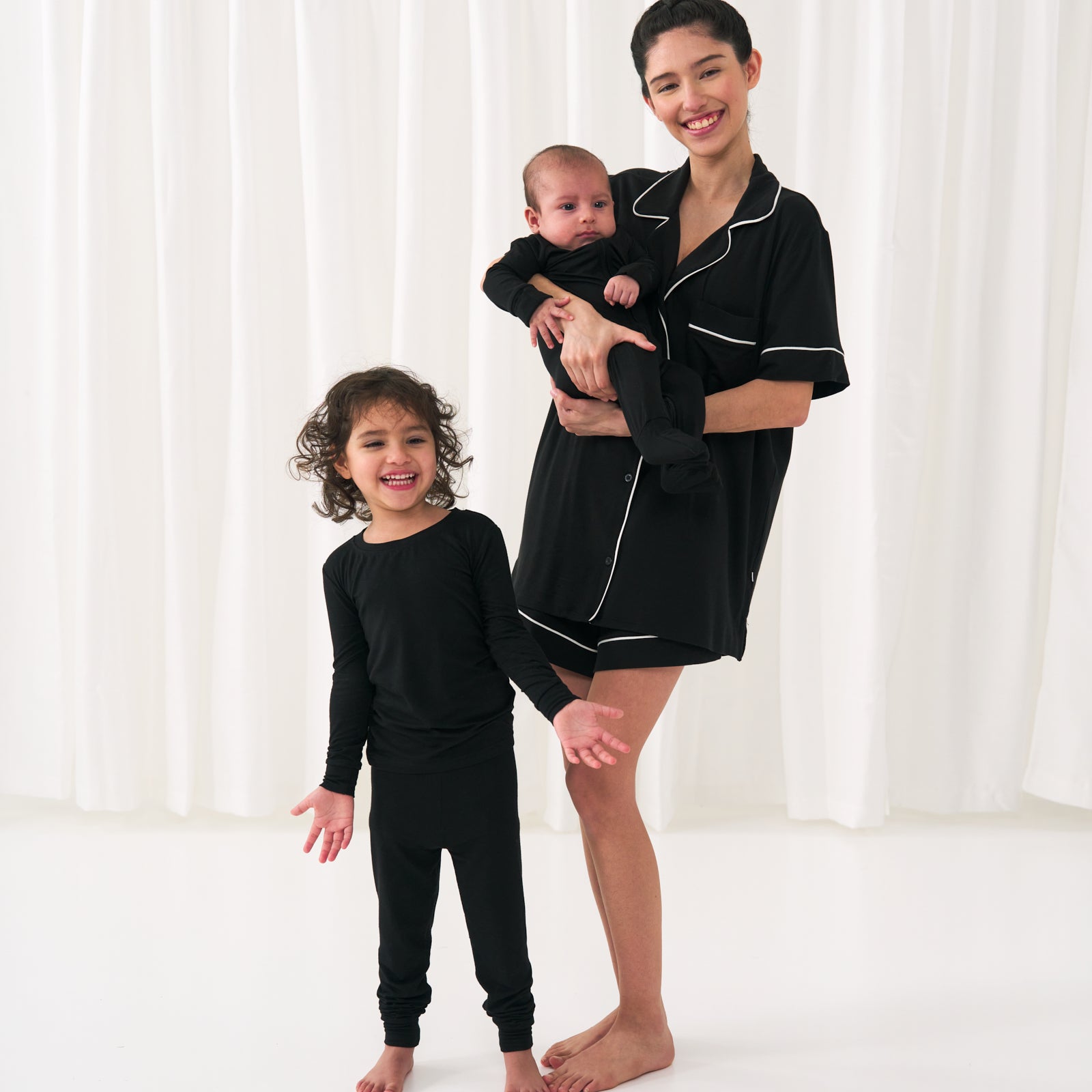 Image of a woman with her two children. Mom is wearing a solid Black women's short sleeve and shorts pajama set. Her kids are wearing Solid Black pjs in two piece and zippy styles