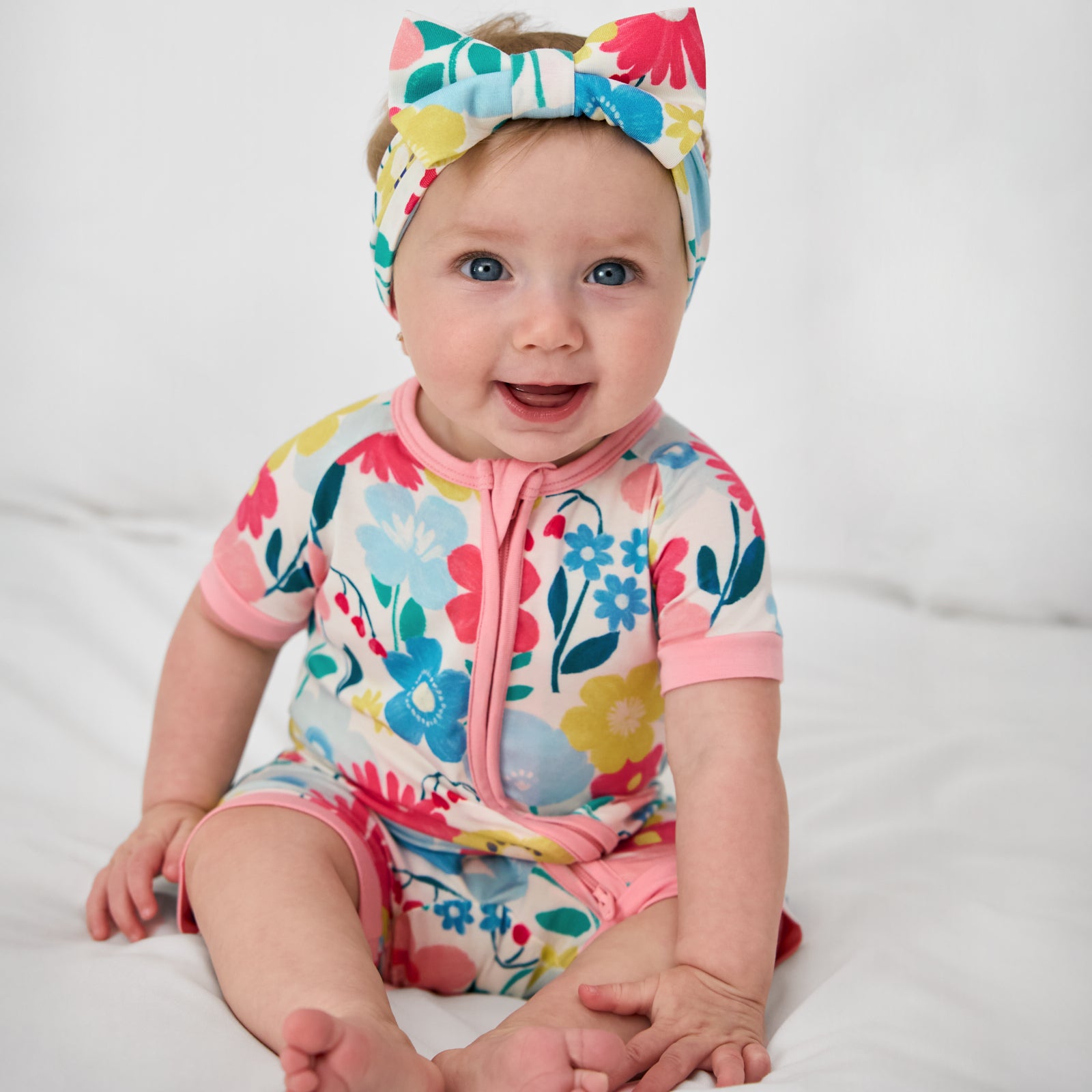 Sitting baby wearing the Midsummer Meadow Shorty Zippy with a matching luxe bow headband
