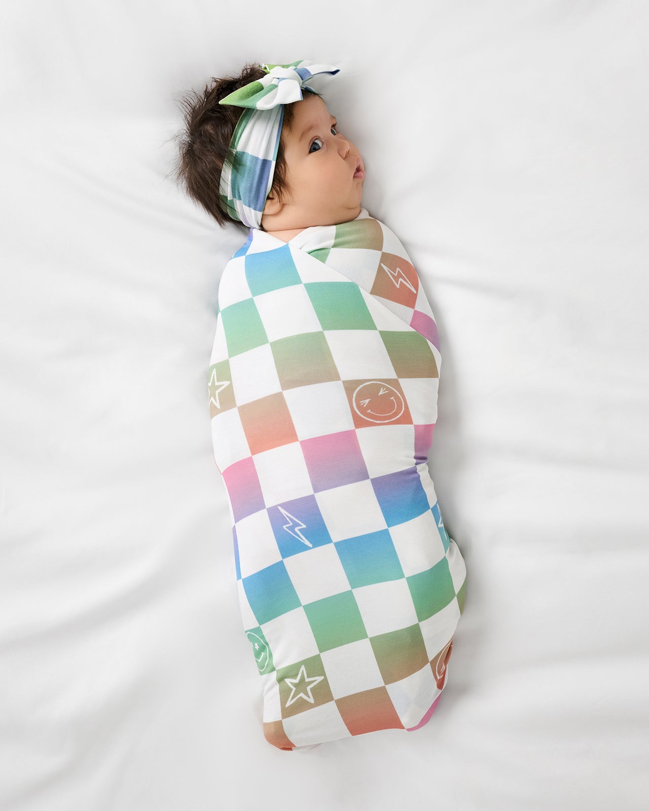 Aerial view of baby wearing the Ombre Cheery Checks Swaddle & Luxe Bow Headband Set