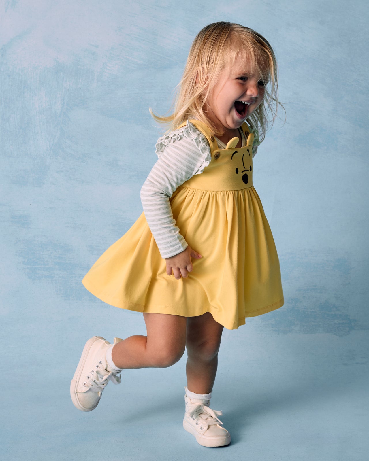 Toddler wearing the Light Heather Gray Stripe Flutter Bodysuit layered under the Winnie the Pooh overall dress on a lue background