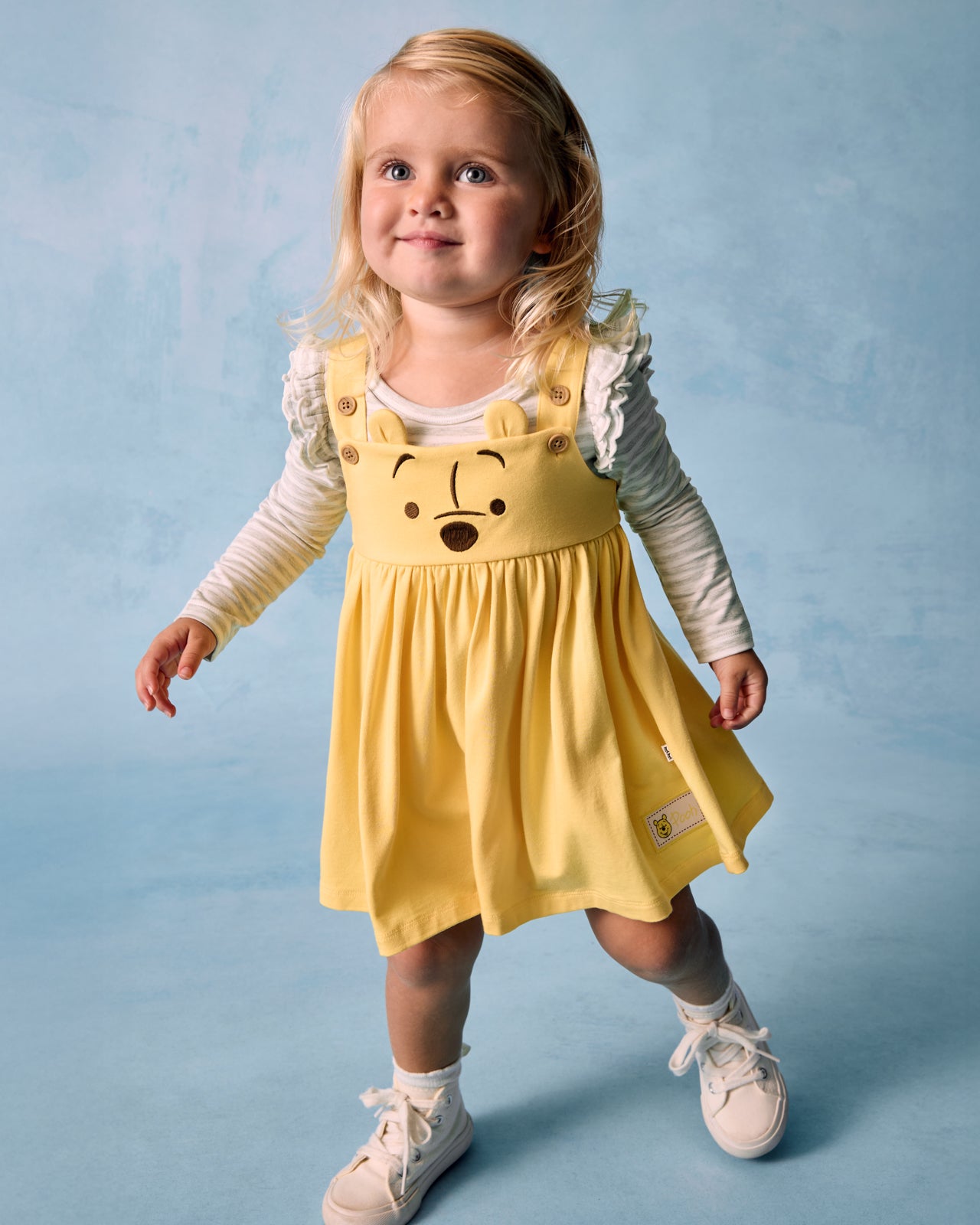 Girl wearing the Disney Winnie The Pooh Overall Dress on a blue background