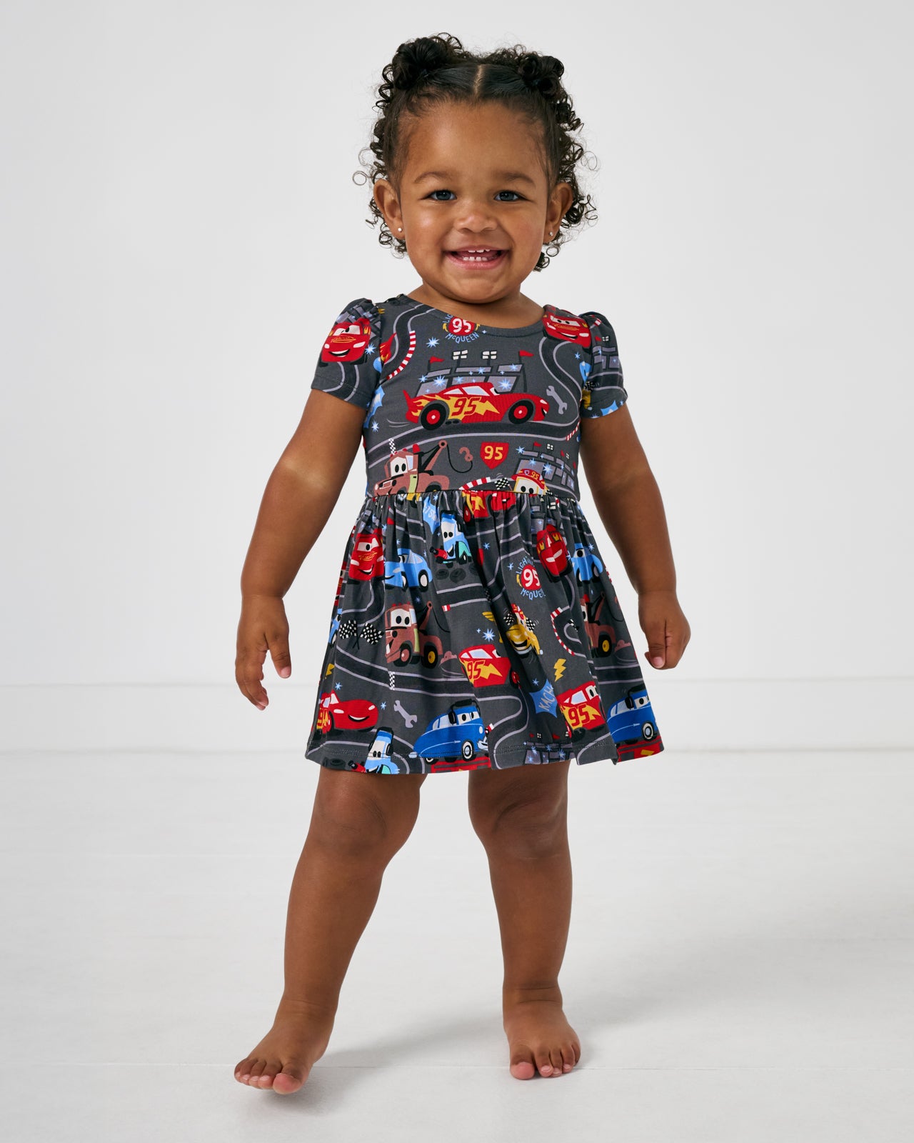 Standing toddler wearing the Disney & Pixar Piston Cup Bow Back Skater Dress with Bodysuit