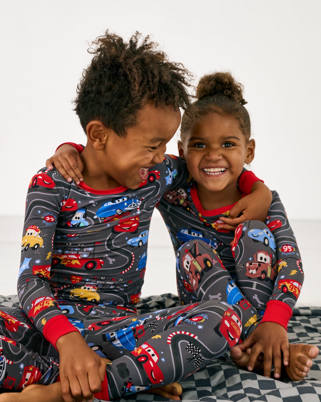 Two children wearing matching Disney & Pixar Piston Cup Two-Piece Pajama Sets sitting on the checkered print blanket
