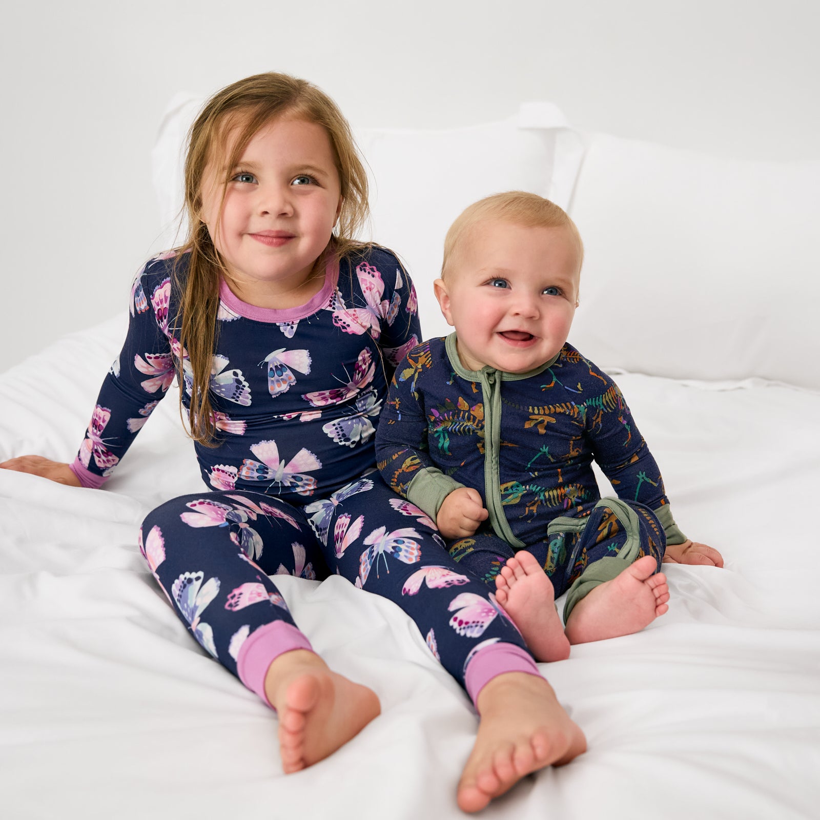 Boy and girl sitting on a bed wearing dinosaur and butterfly themed pjs