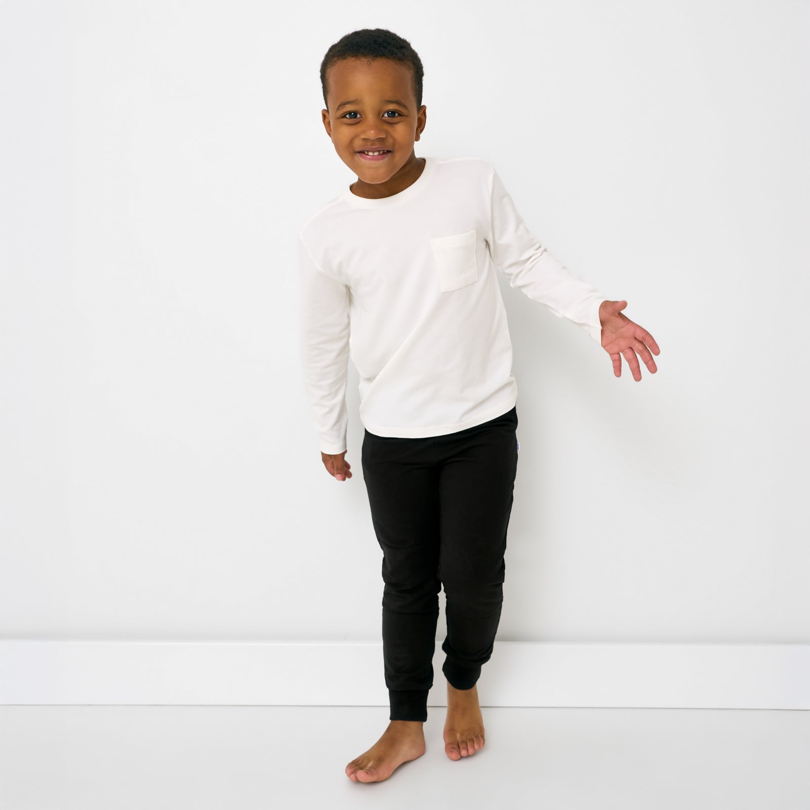 Standing child wearing the Soft White Long Sleeve Relaxed Pocket Tee with black joggers