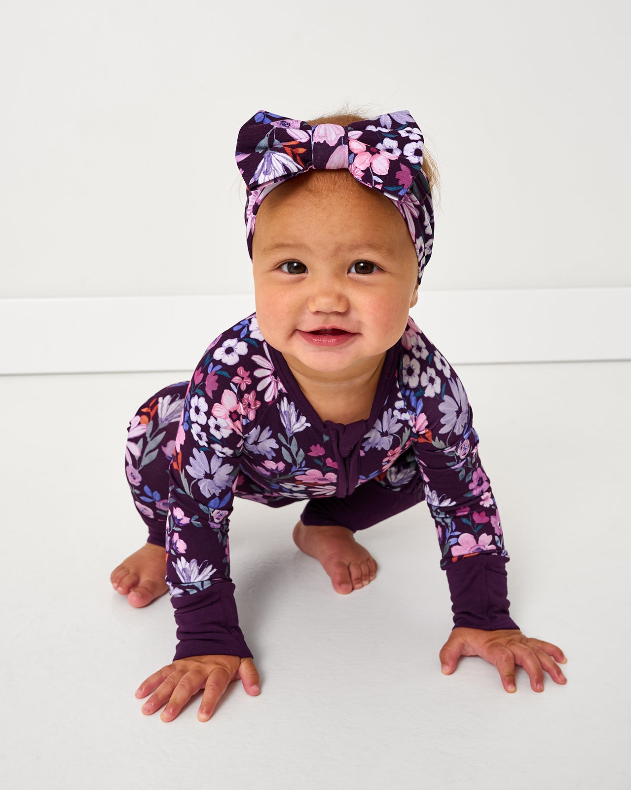 Crouching baby wearing the Violet Meadow Luxe Bow Headband with floral zipper baby pajamas