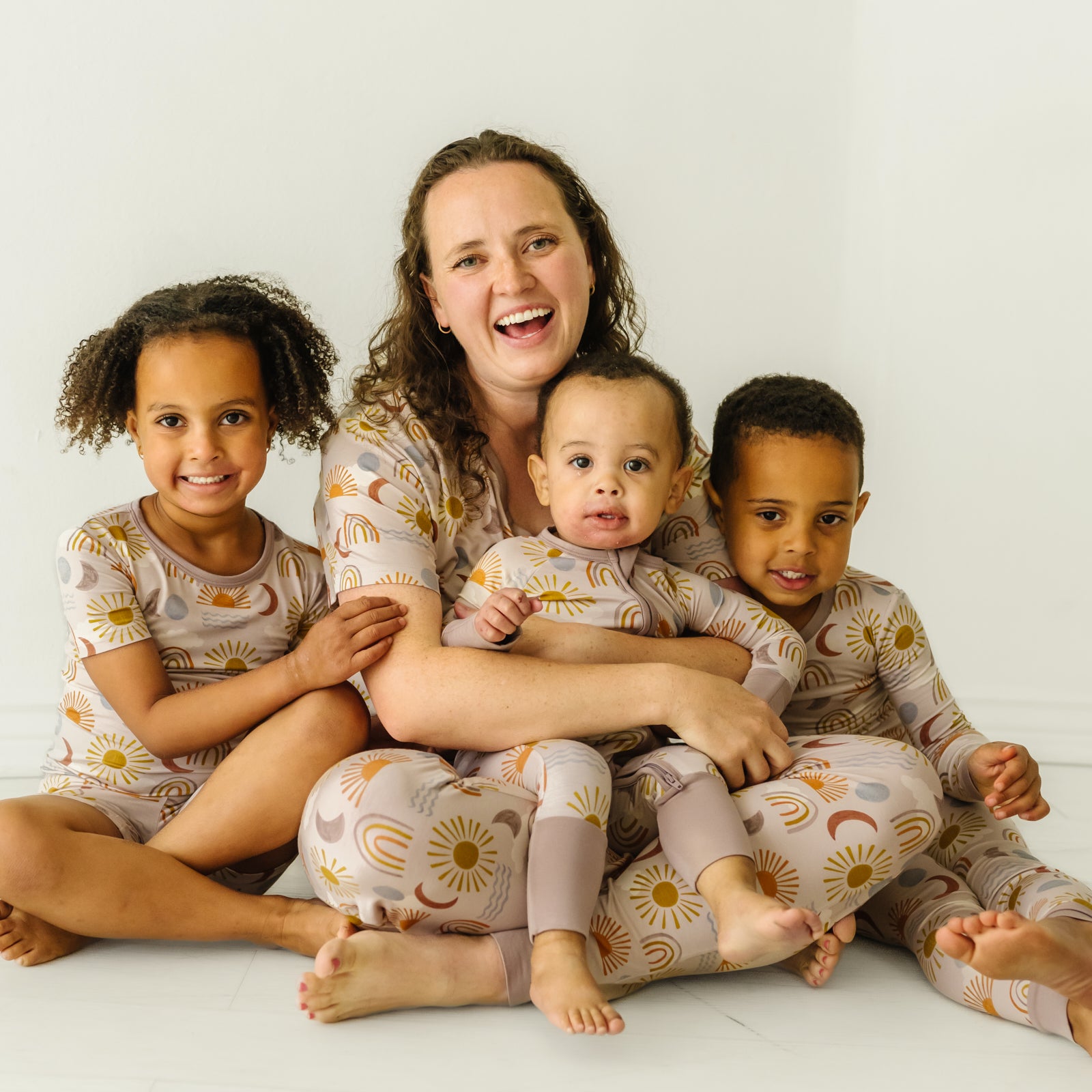 Mother sitting with her three children wearing matching Desert Sunrise pajamas. Mom is wearing Desert Sunrise women's short sleeve pj top and matching women's pj pants. Kids are wearing Desert Sunrise pjs in two piece and zippy styles