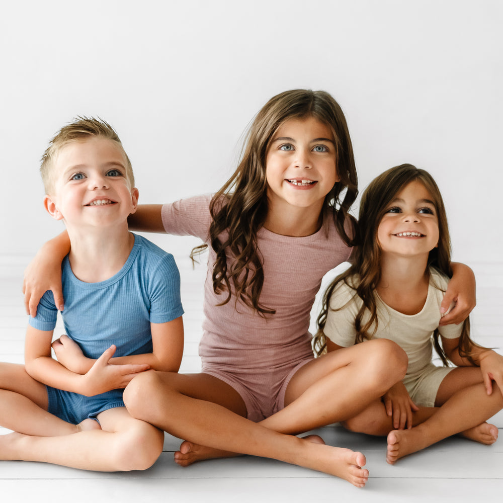 Three children sitting together wearing coordinating Ribbed two piece short sleeve and shorts pajama sets in Heather Blue, Heather Mauve, and Heather Oatmeal