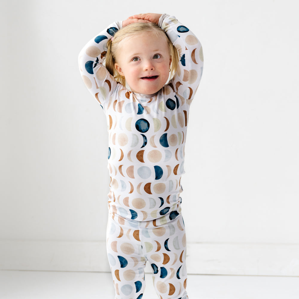 Image of a toddler in long sleeve top and pajama pants set in Luna Neutral print. This print features phases of the moon in the sweetest shades of creams, tans, and navy watercolor in an all over repeat pattern.