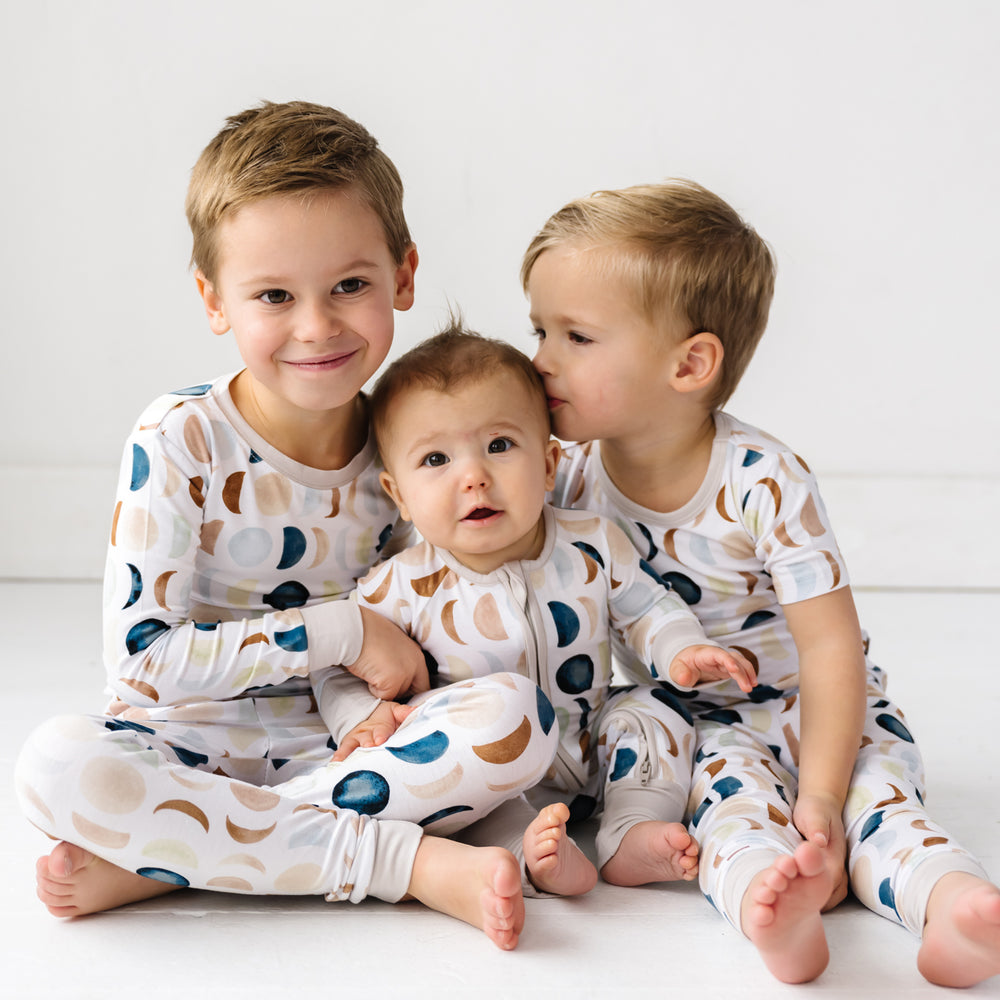 Image of three young children in matching Luna Neutral printed pajamas. This print features phases of the moon in the sweetest shades of creams, tans, and navy watercolor in an all over repeat pattern.