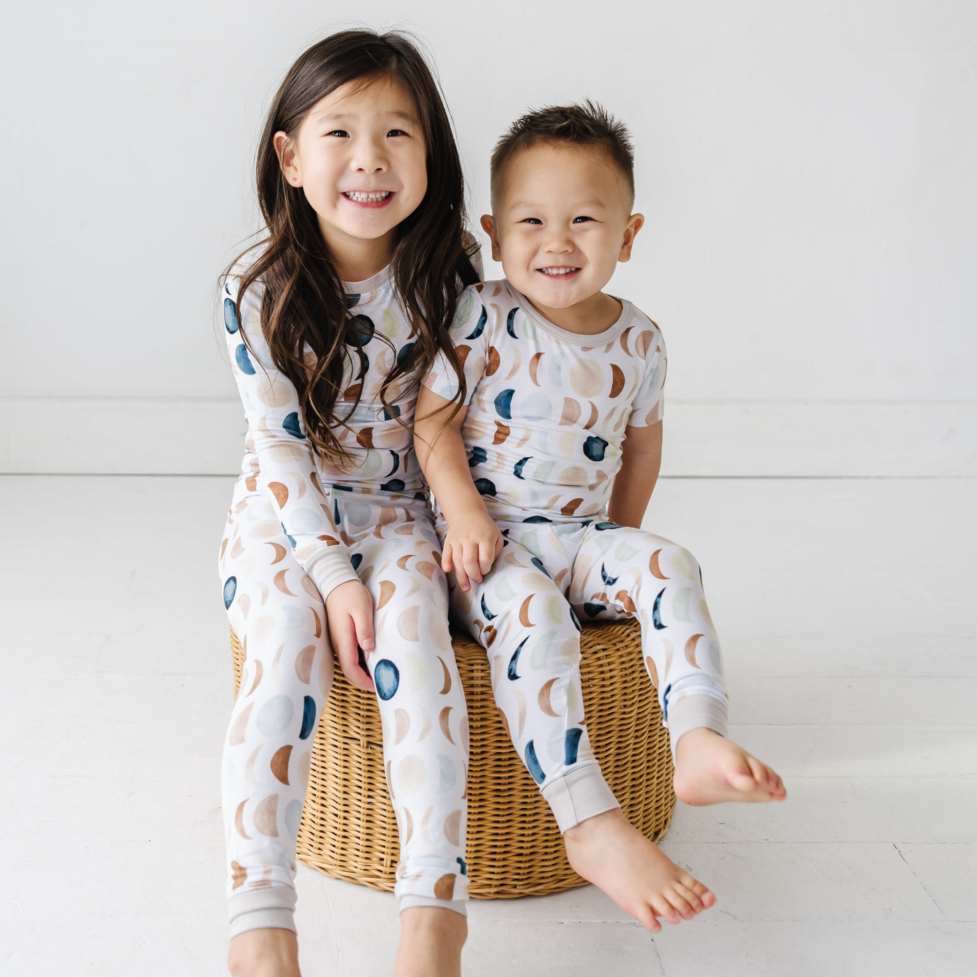 Image of two toddlers in pajama sets in Luna Neutral print. This print features phases of the moon in the sweetest shades of creams, tans, and navy watercolor in an all over repeat pattern.