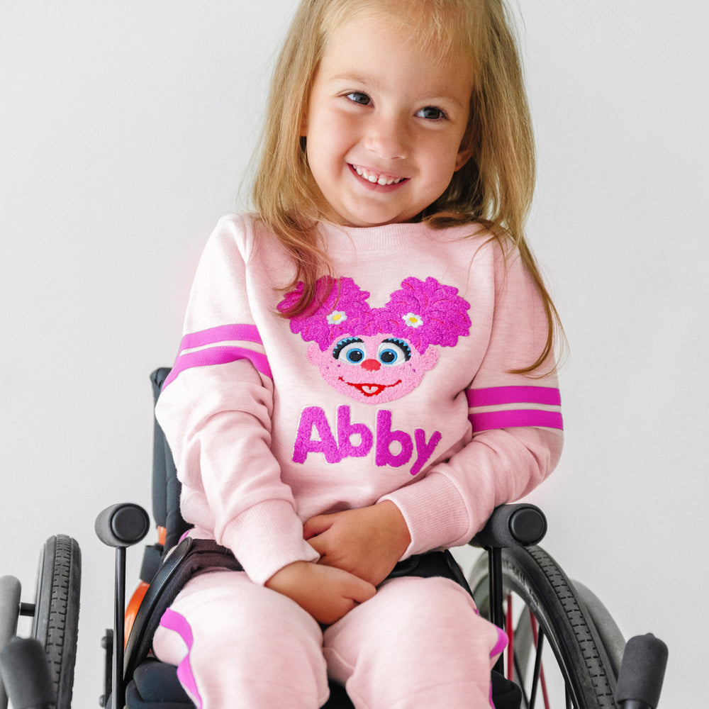 Close up image of a child in a wheelchair wearing a Sesame Street Abby crewneck sweatshirt and jogger set