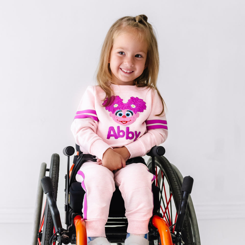 Child in a wheelchair wearing a Sesame Street Abby crewneck sweatshirt and jogger set