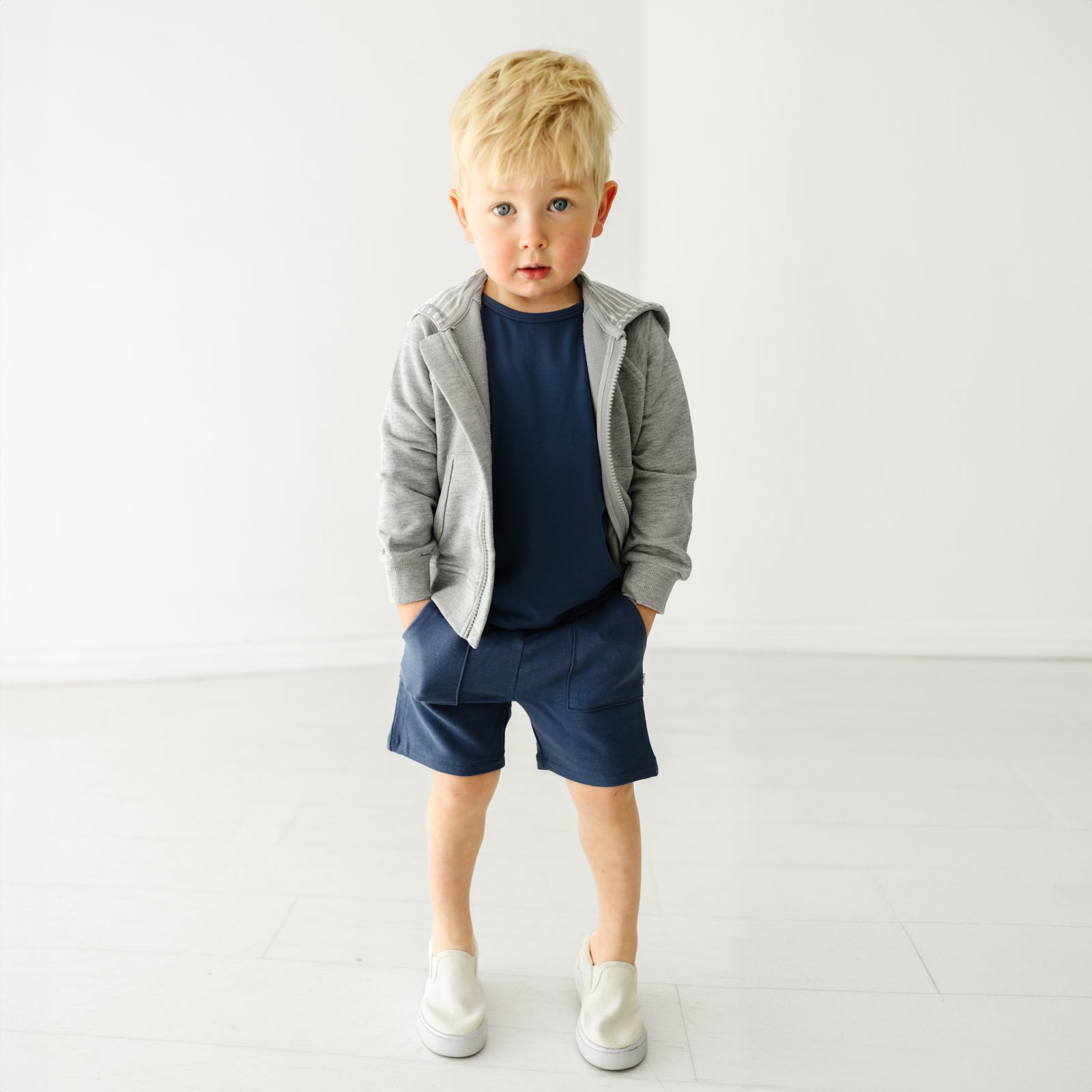 Child with their hands in their pockets wearing Vintage Navy Shorts, Classic Navy Tee, and Heather Gray Zip Hoodie.