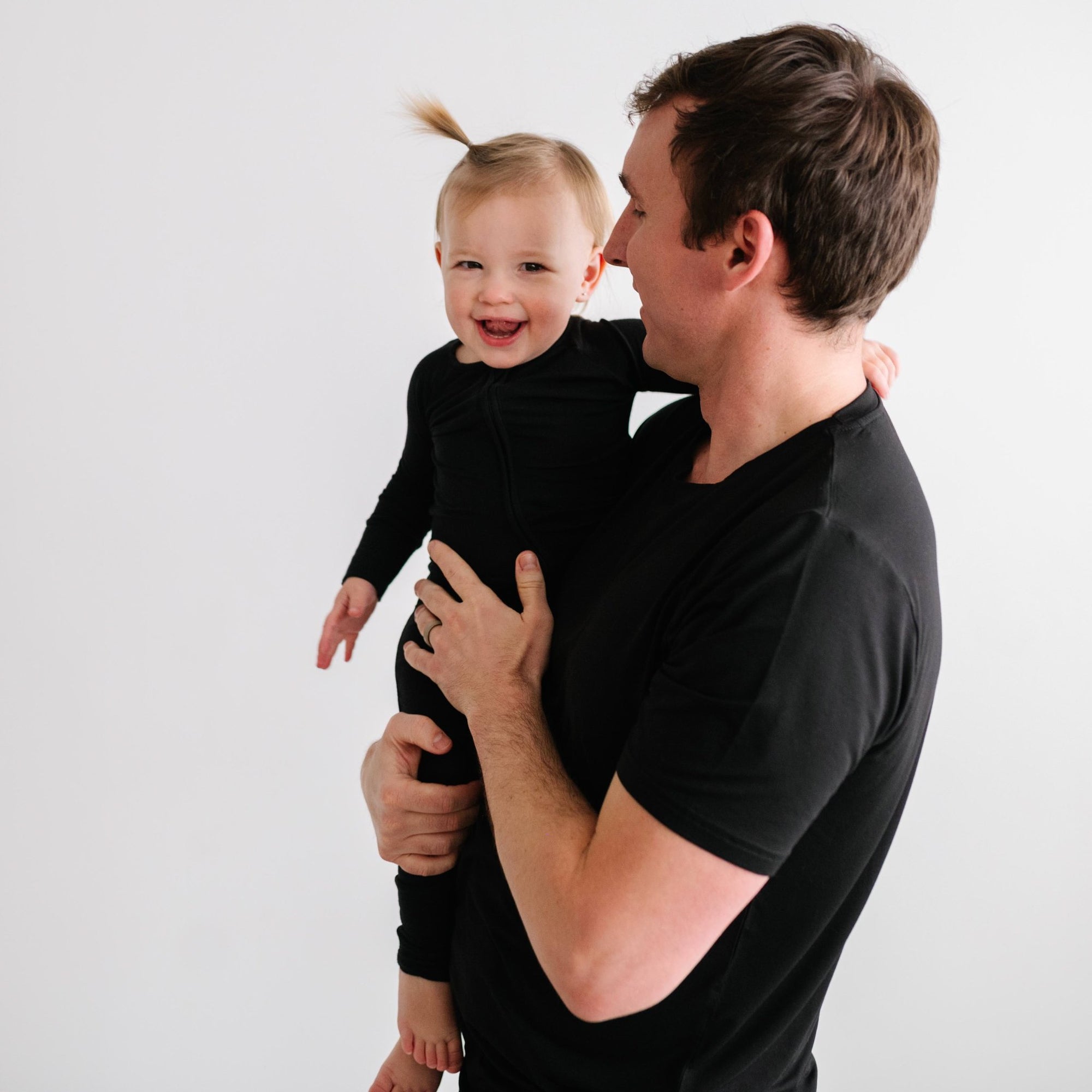 Image of dad holding his daughter. They are both shown wearing matching solid black pajamas. The dad is shown wearing a solid black short sleeve pajama top, and the daughter is shown wearing a solid black zip up romper.