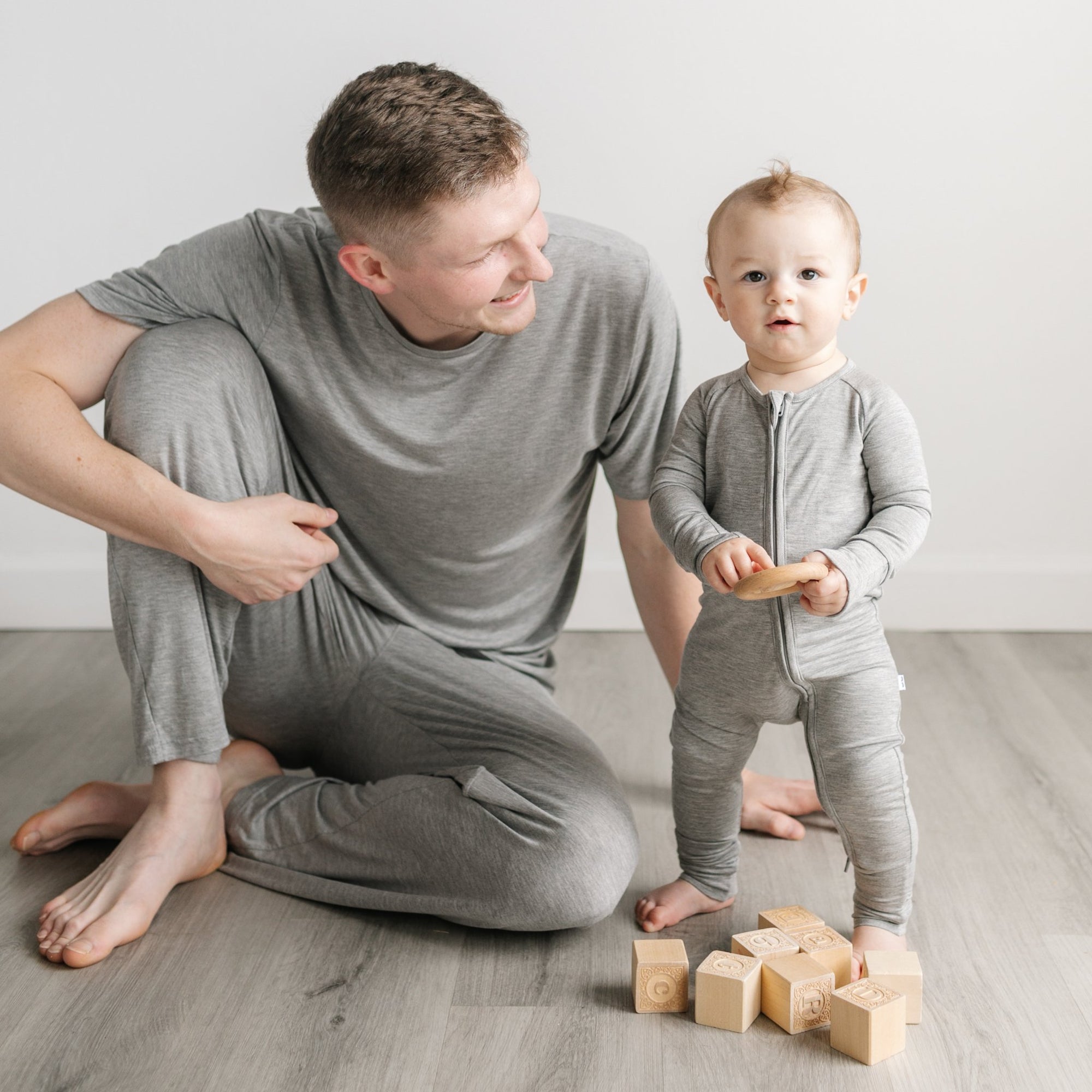 Image of a dad and son playing with wooden building blocks. The dad is shown wearing a short sleeve pajama top and pajama pants in heather gray, while the son is shown wearing a zip up romper in heather gray.