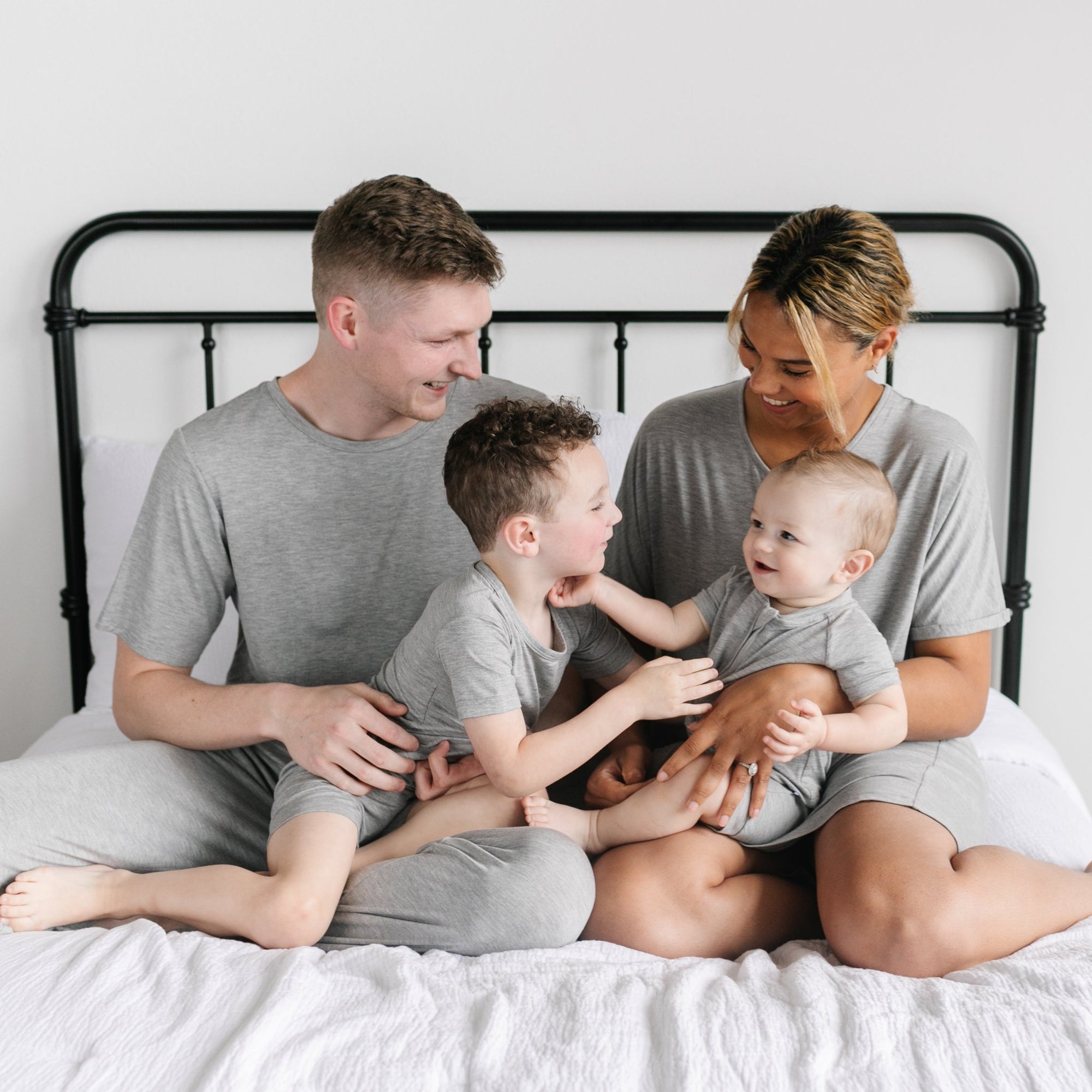 Image of family of four all sitting on a bed. They are shown wearing matching solid heather gray pajamas. The dad is shown wearing a short sleeve pj top and pants, the mom is shown wearing a short sleeve caftan gown, one son is shown wearing a short sleeve and shorts pj set, while the other son is shown wearing a short sleeve and shorts zip up romper.