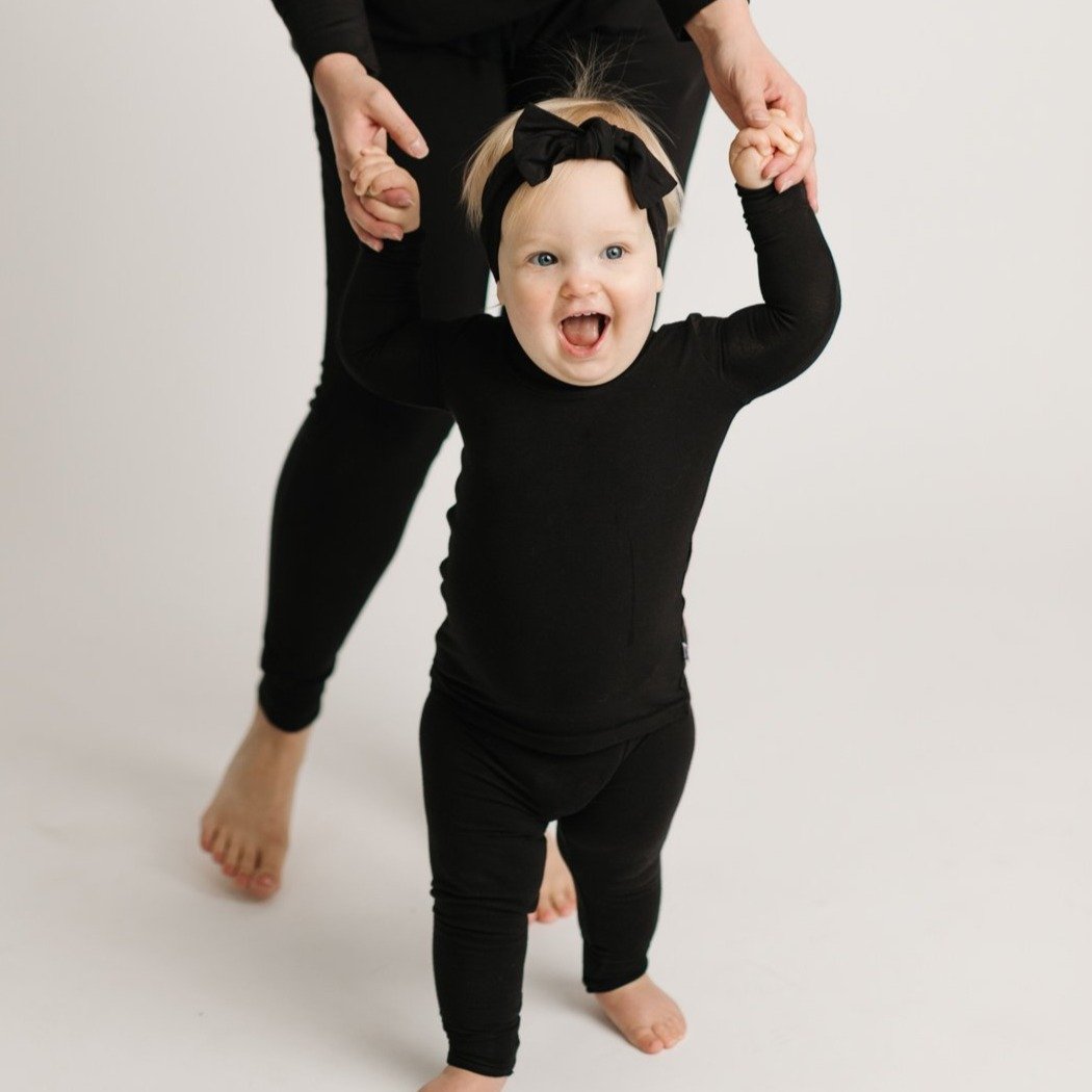 Baby girl wearing solid black two-piece pajama set with matching solid black bow headband.