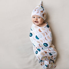 Image of an infant in a swaddle and knotted hat set in Luna Neutral print. This print features phases of the moon in the sweetest shades of creams, tans, and navy watercolor in an all over repeat pattern.