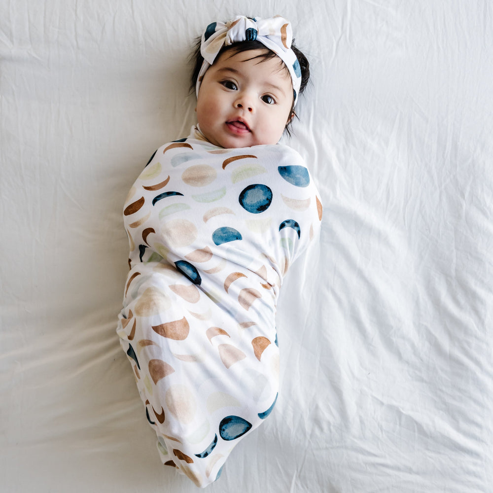 Image of an infant in a swaddle and bow headband set in Luna Neutral print. This print features phases of the moon in the sweetest shades of creams, tans, and navy watercolor in an all over repeat pattern.