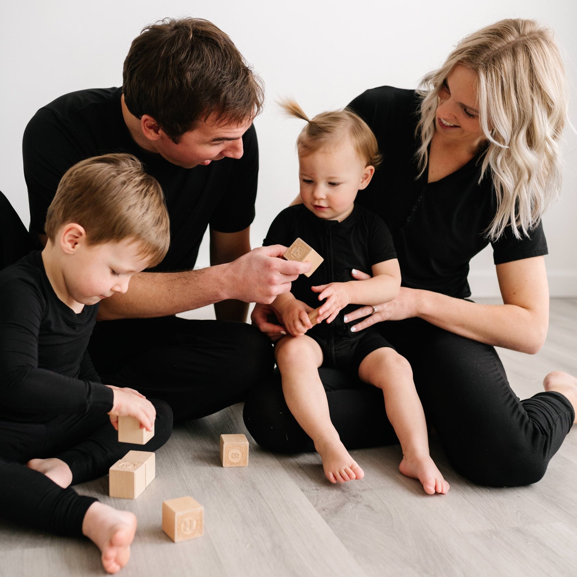 Image of family of 4 all sitting down on the floor and playing with wooden building blocks. They are all shown wearing matching solid black pajamas. The mom and dad are both shown wearing solid black short sleeve pj tops with matching solid black pj pants