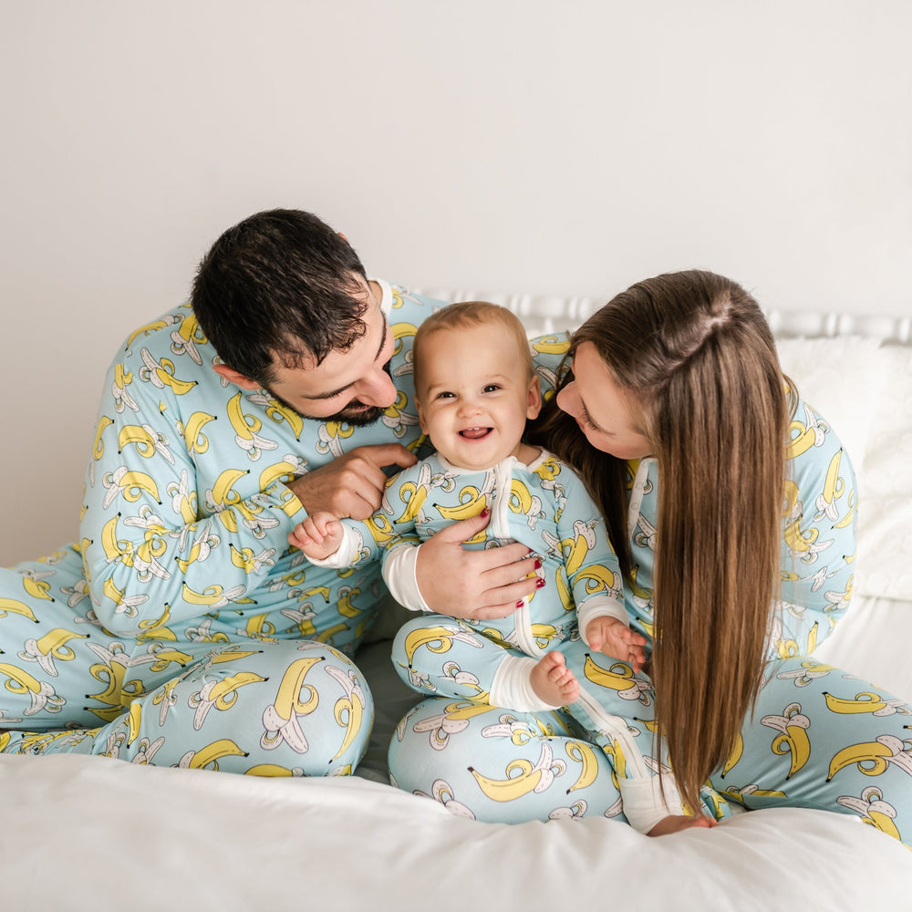 Family of three all wearing matching banana printed pajamas. The mom and dad pajama sets have a light blue background with white trim accents and pops of yellow coming from the banana print. While the baby boy's zip up romper has a light blue background with white trim accents and pops of yellow coming from the banana print.