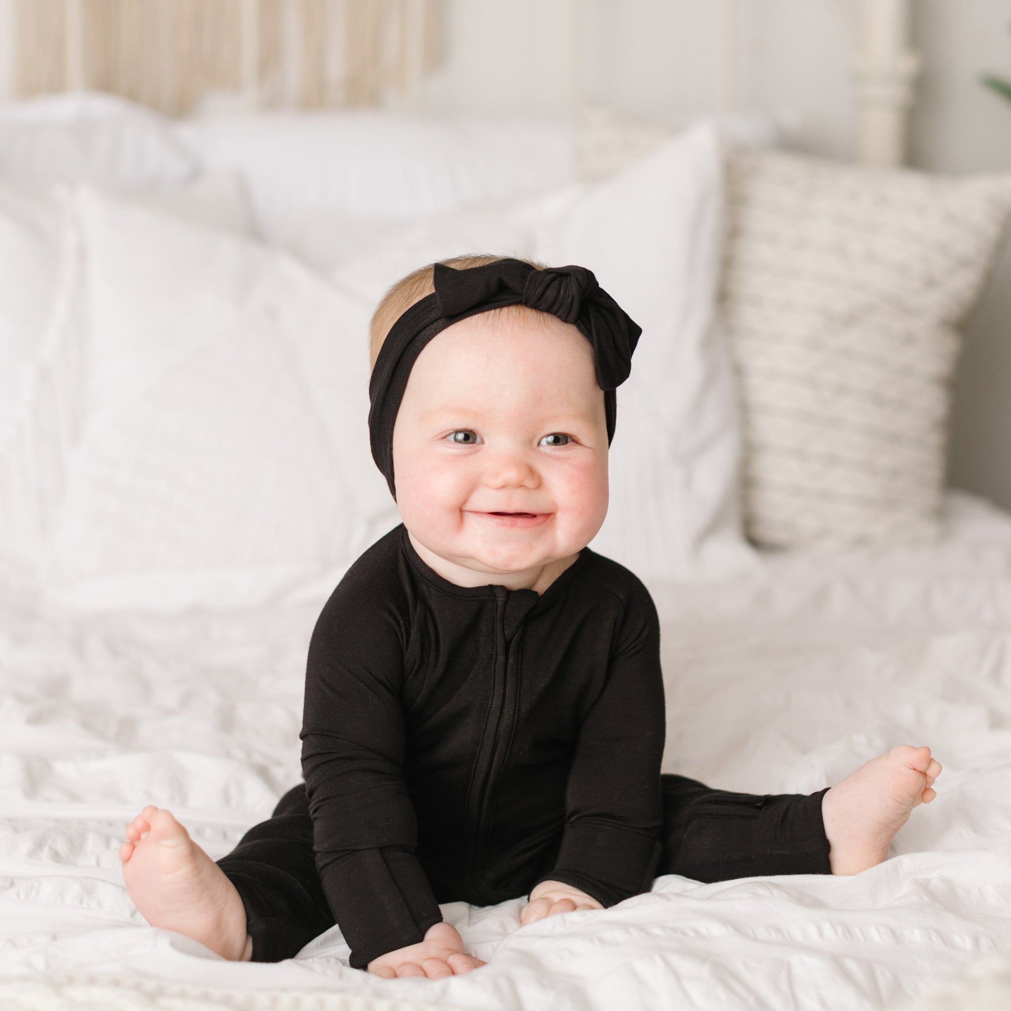 Image of baby girl sitting on a bed wearing solid black zip up romper with matching solid black bow headband.