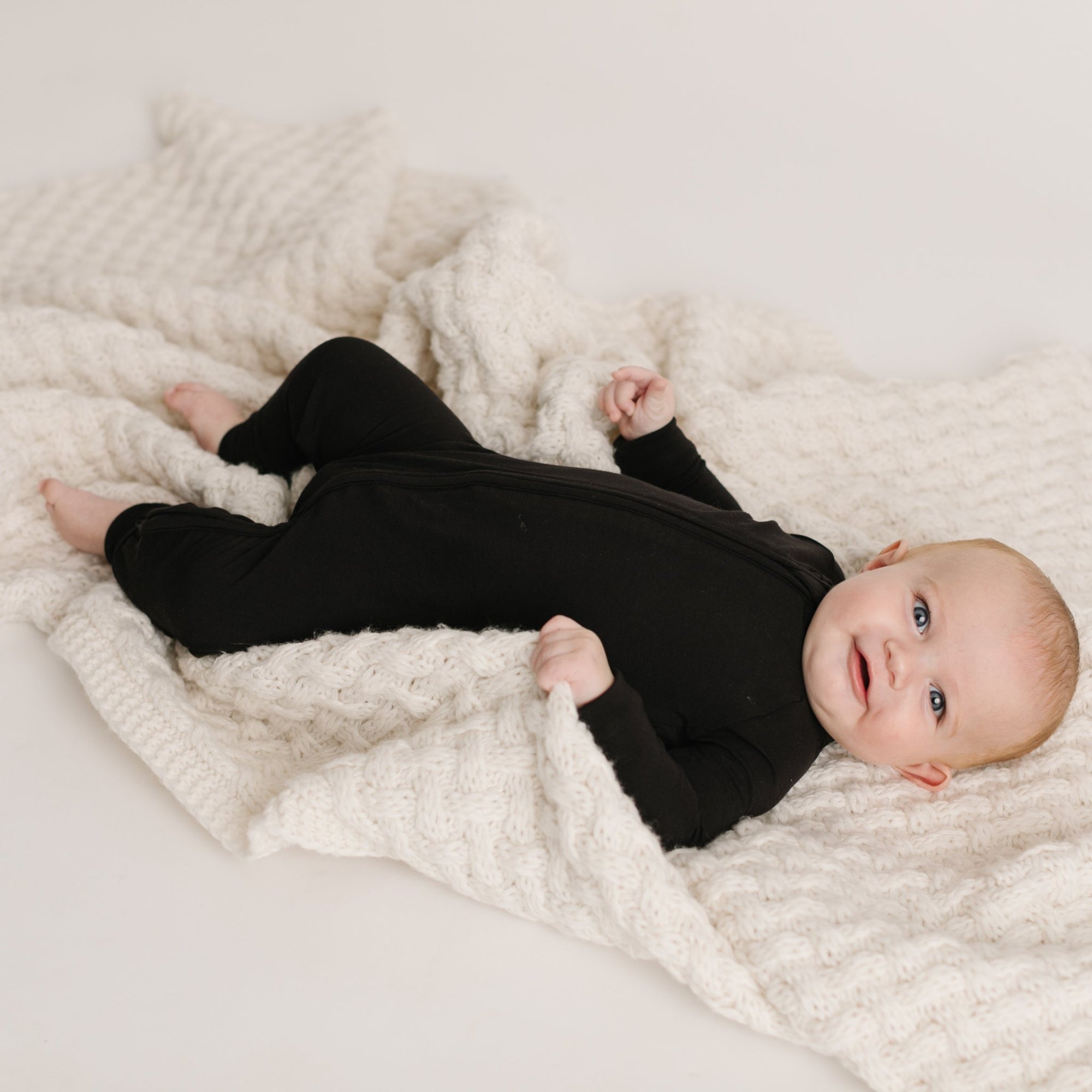 Image of baby boy laying down on a blanket wearing solid black zip up romper.