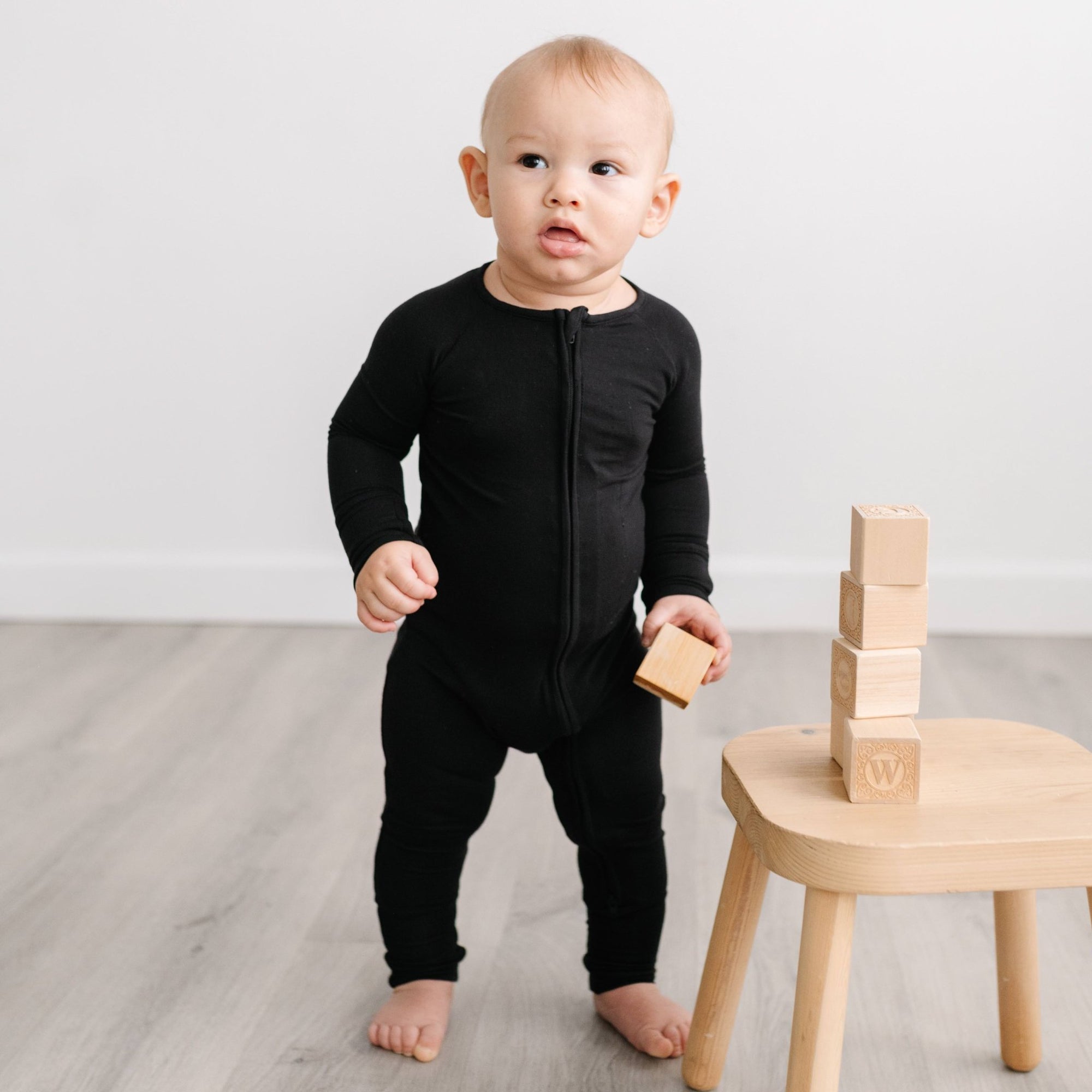 Image of infant boy holding a wooden building block in his hand. He is shown wearing a solid black zip up romper.