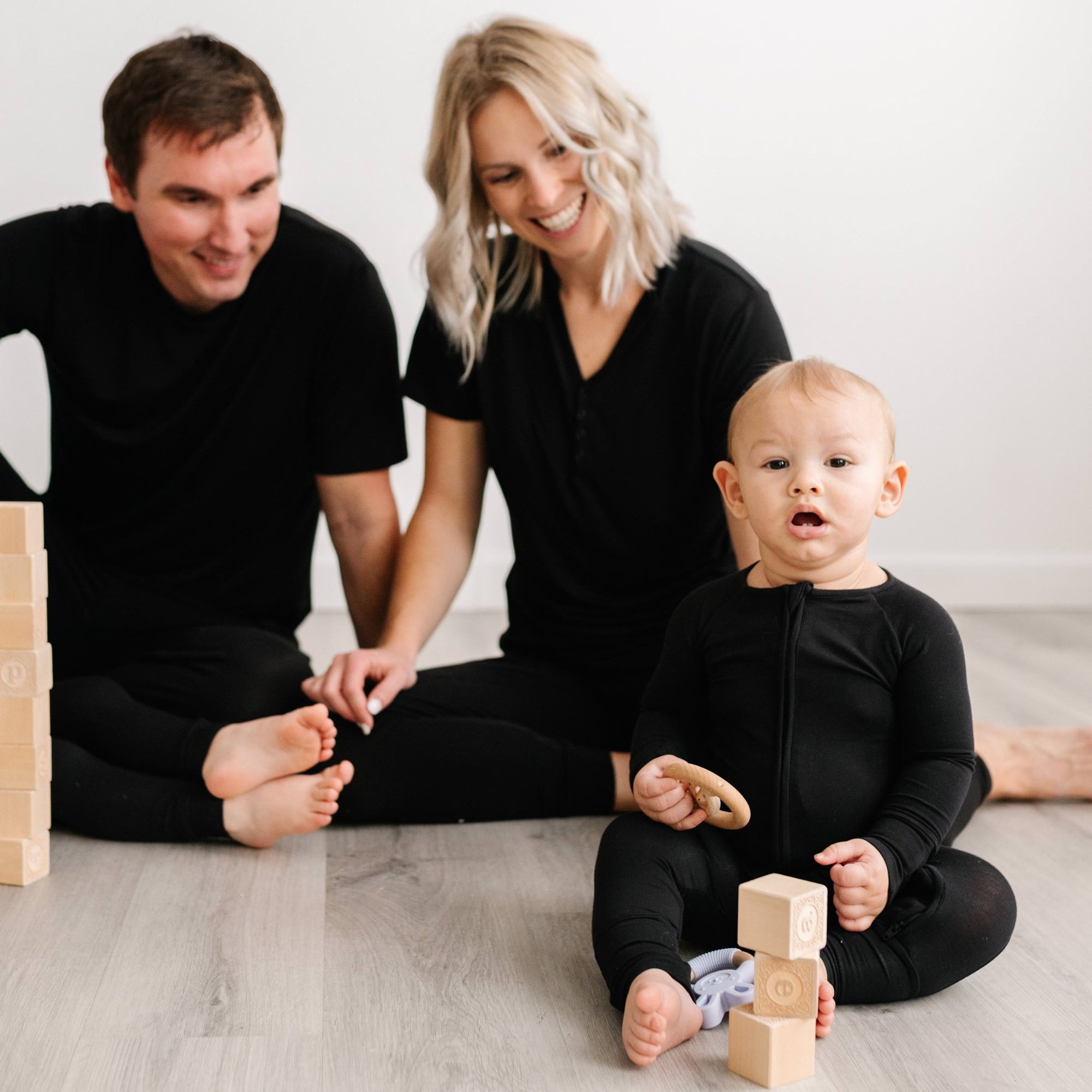 Image of family of 3 sitting on the floor and playing with wooden building blacks. They are all shown wearing matching solid black pajamas. The mom and dad are both shown wearing solid black short sleeve pj tops with matching solid black pj pants and the
