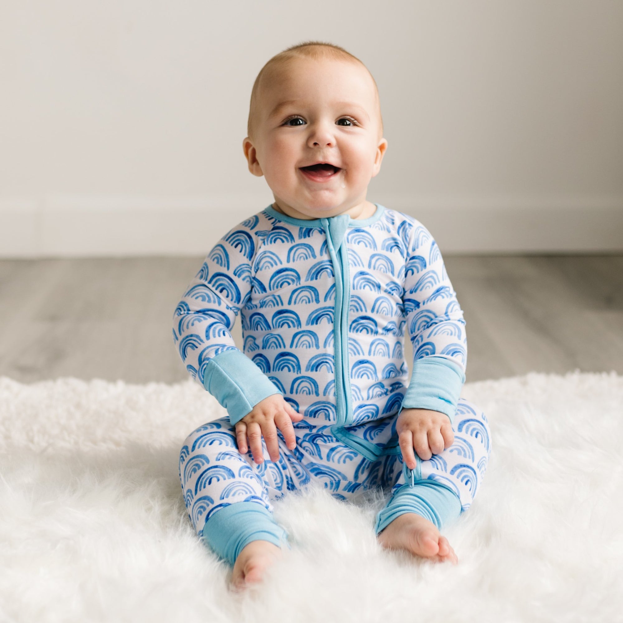 Image of baby boy wearing blue rainbow printed zip up romper. This print sits on a white background with shades of blue rainbows and sky blue trim details.