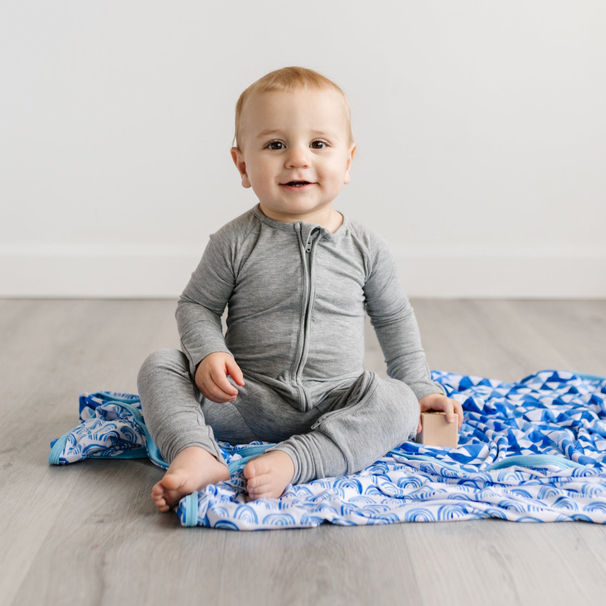 Image of baby boy sitting on top of blue rainbows printed blanket. He is shown wearing zip up romper in heather gray.