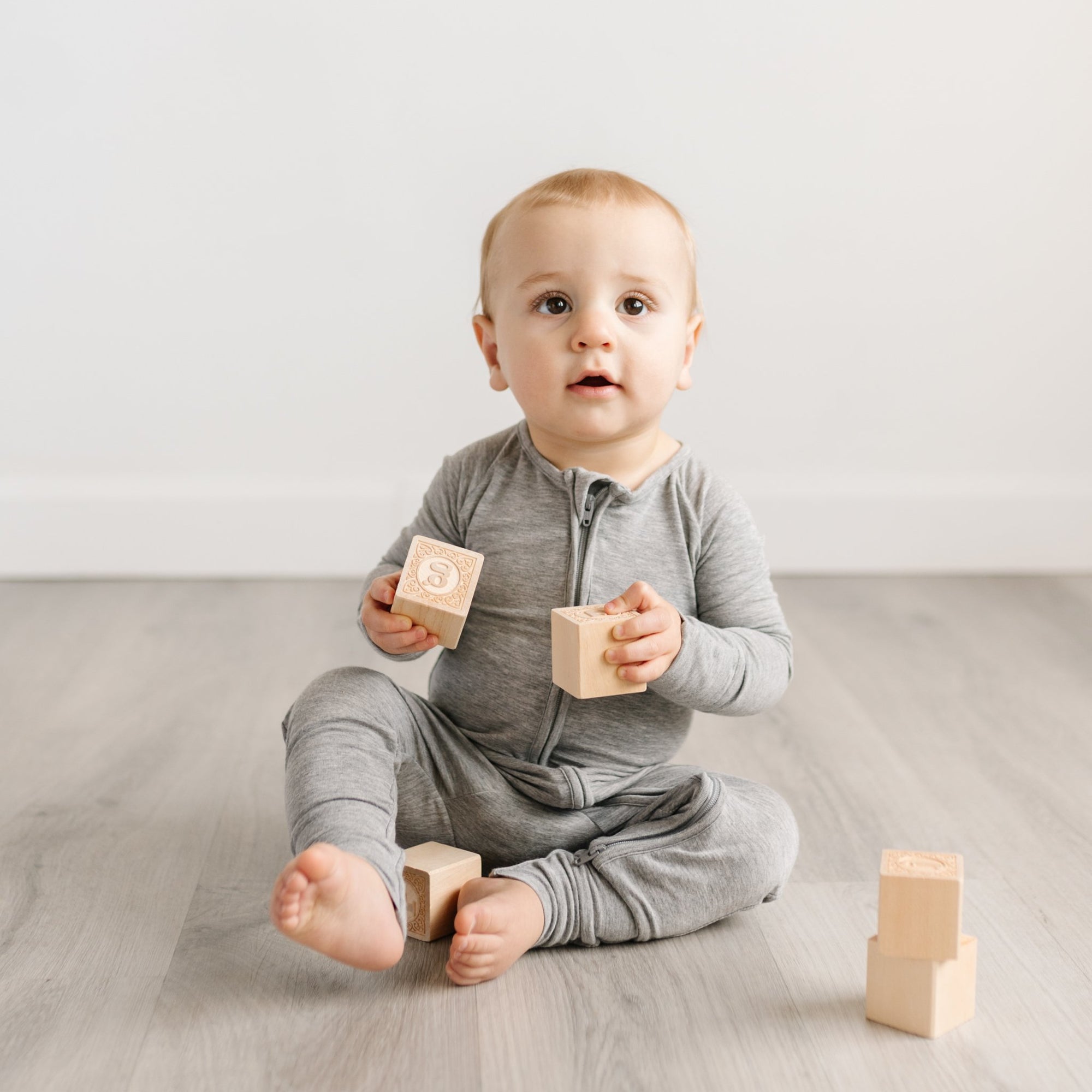 Image of baby boy sitting down and playing with wooden building blocks. He is shown wearing zip up romper in heather gray.