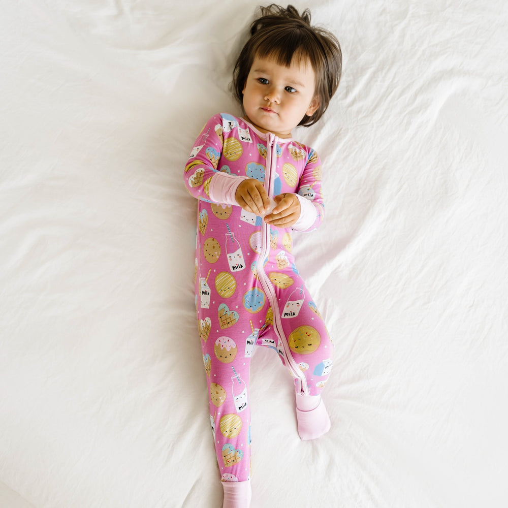 Image of toddler girl wearing a zip up romper in cookies and milk print. This print features milk cartons, colorful sprinkled cookies, and chocolate chip cookies that sit upon a pink background with light pink trim.