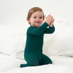 A young child with light brown hair wearing a green onesie is sitting on a white bed, clapping their hands with a joyful expression.