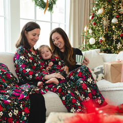 Three people are sitting on a couch in matching floral pajamas. A Christmas tree and wreath are visible in the background. The adult on the left is holding a child, while the adult on the right is holding a striped mug.