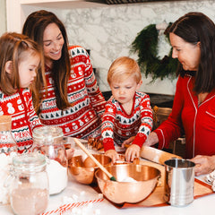 A family in matching Christmas pajamas bakes together in a kitchen. Two children and two women are seen preparing dough with various ingredients and utensils on the counter. A festive wreath hangs in the background.