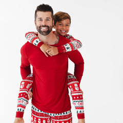 A man with a beard, dressed in red Christmas-themed pajamas, smiles while giving a piggyback ride to a child in matching pajamas. Both are looking at the camera against a plain white background.