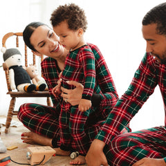 A family of three, wearing matching red and green plaid pajamas, sits together. The mother holds a young child who stands in the center. A chair with two soft toys and a wooden toy car are visible in the background.