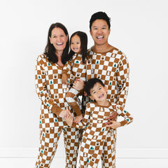 A family of four, two adults and two children, stand together wearing matching brown and white checkered pajamas with small illustrated designs. They are all smiling and posing in front of a plain white background.